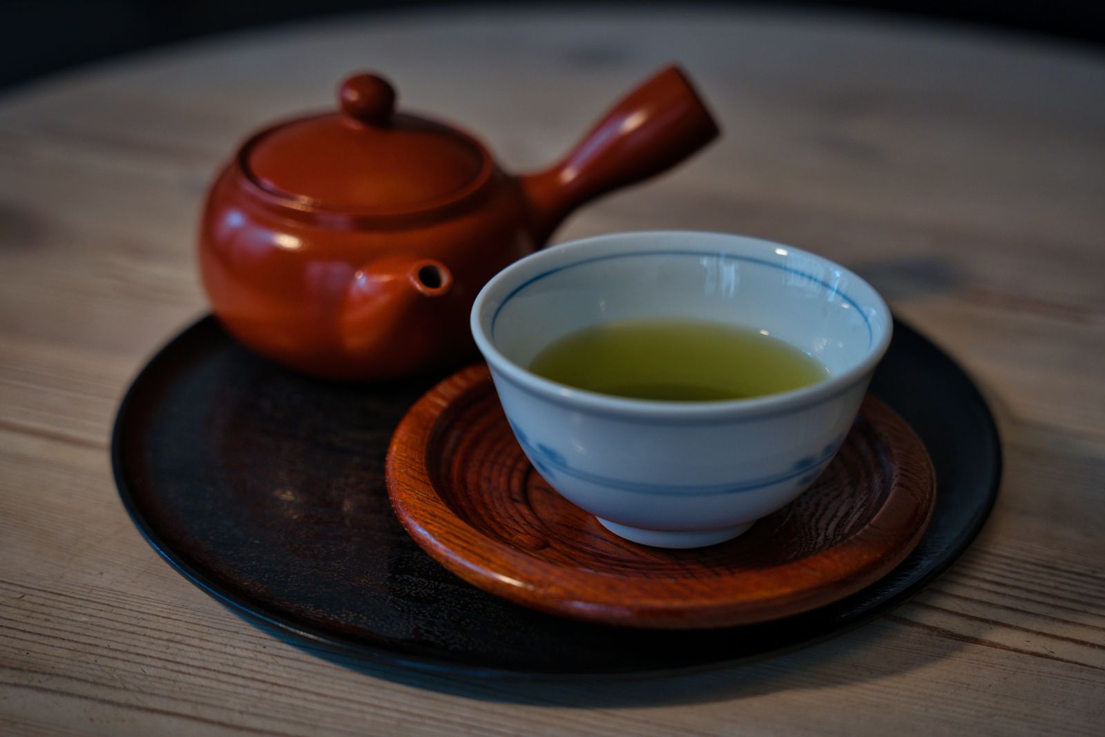 A red-brown kyusu with its side handle in frame, beside a white porcelain cup of pale green tea on a wooden coaster and a black lacquer plate.