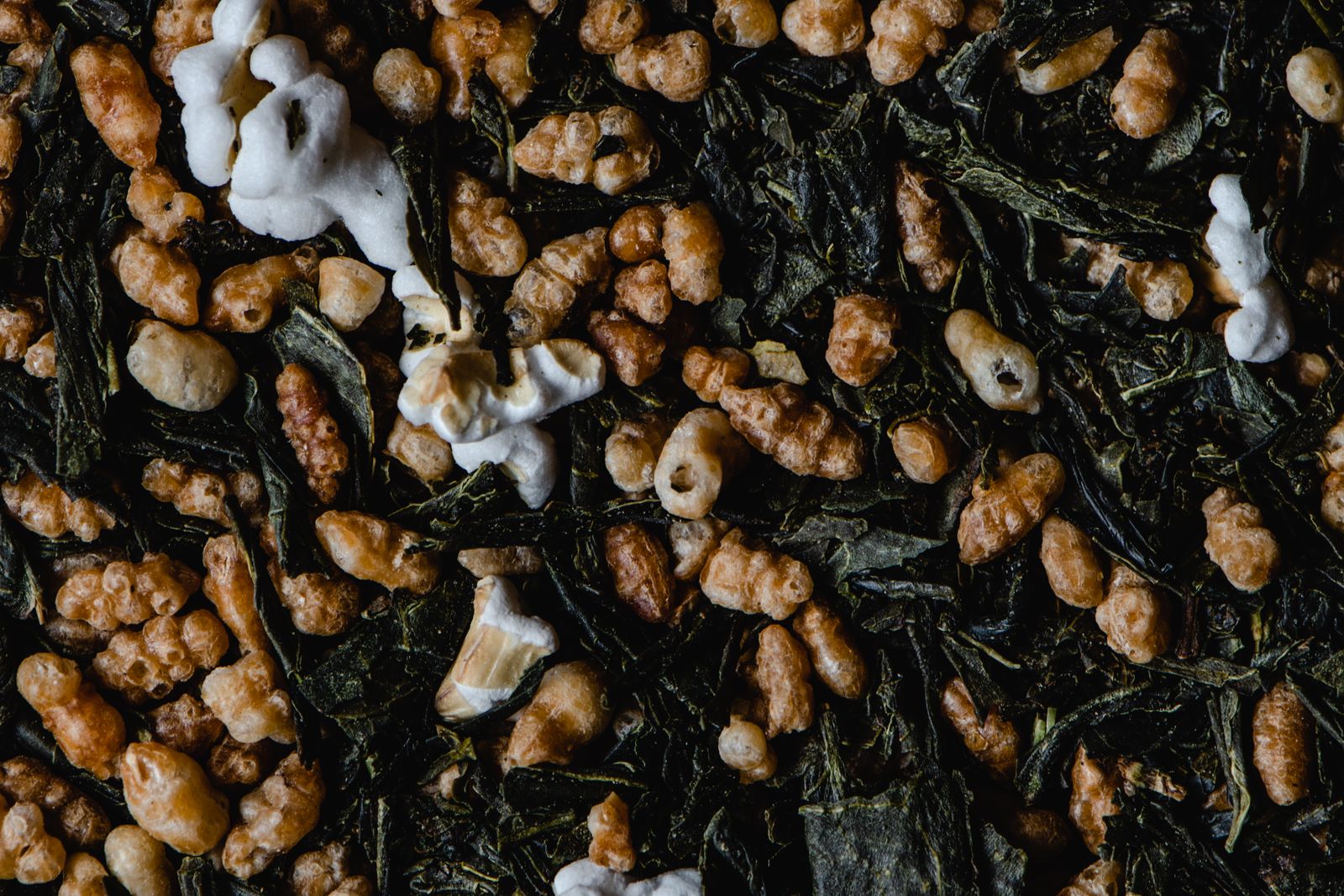 A close view of dark green tea leaf mixed thoroughly with pale golden roasted rice grains, several of them burst open and white.
