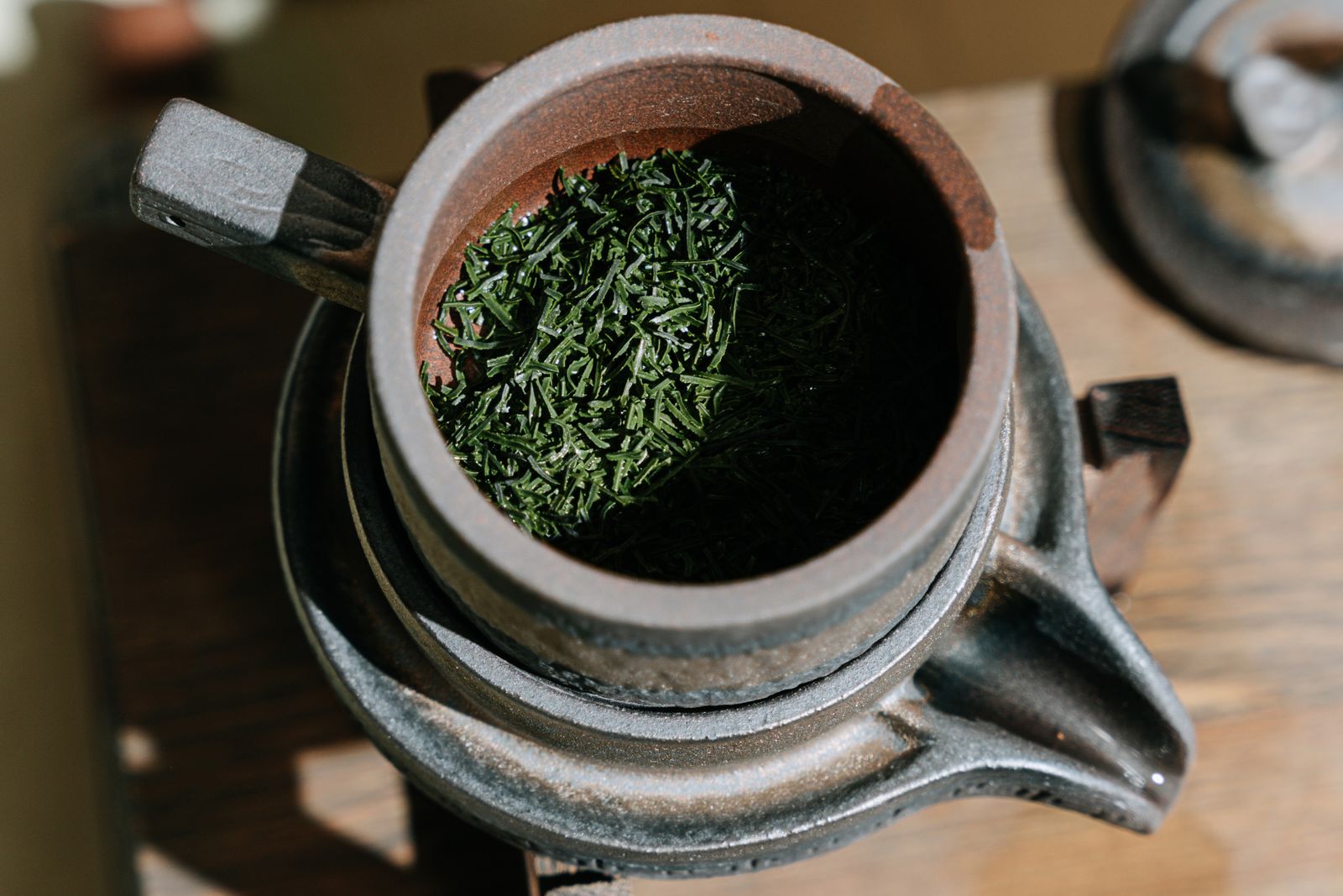 A kyusu seen from above with its side handle and spout in frame, filled with dark green needle-shaped dry leaf, the lid resting beside it.