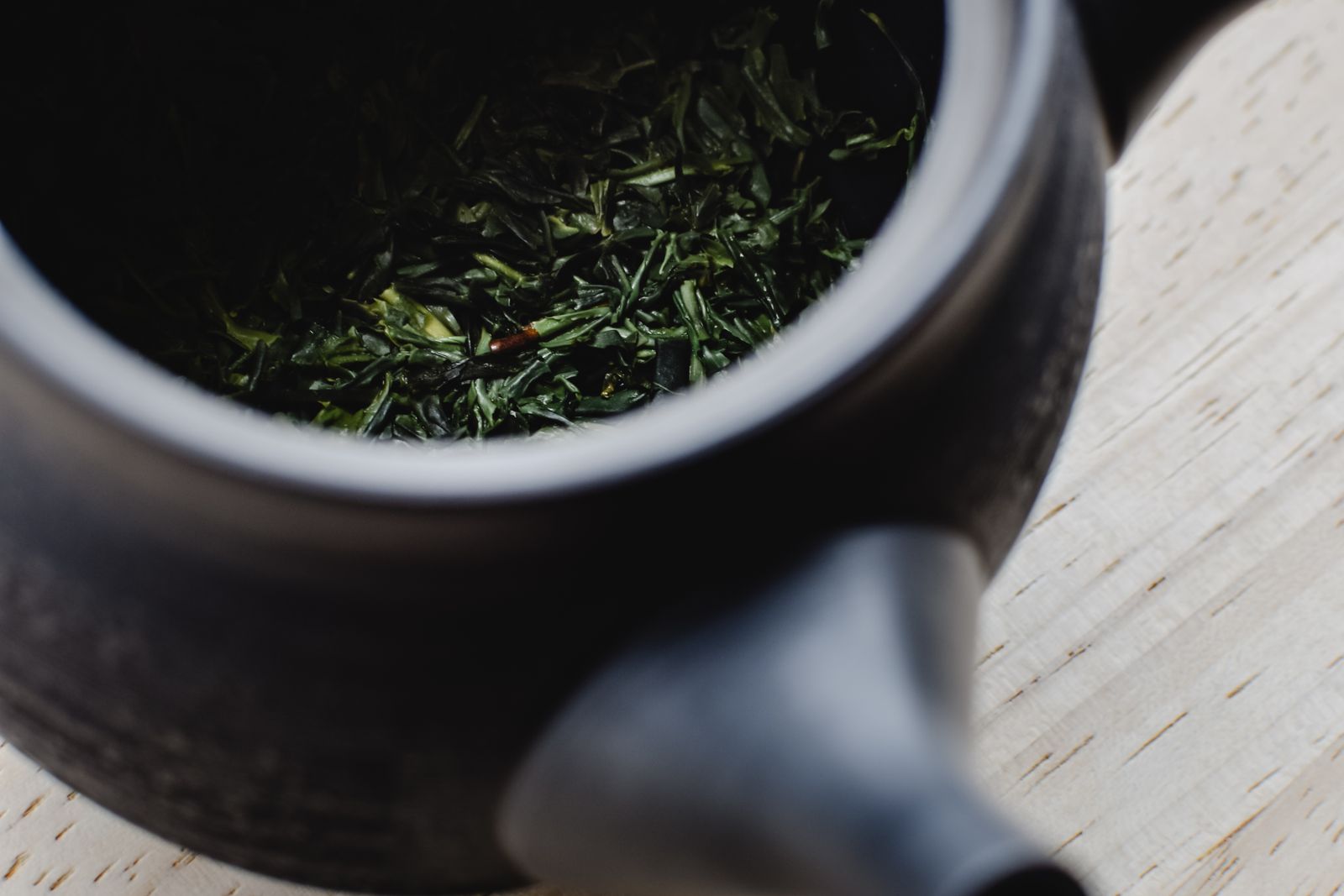 A dark ceramic pot seen from above, holding loose green tea leaf, on a pale wooden surface.