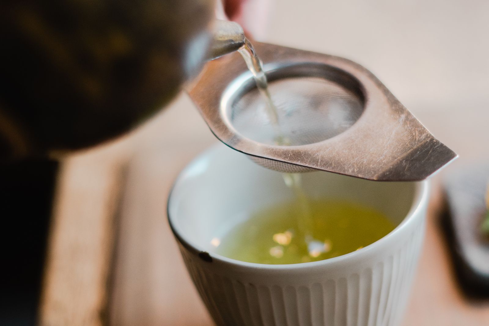 Pale yellow-green tea pouring in a clear stream from a dark pot through a metal mesh strainer into a white ribbed cup.