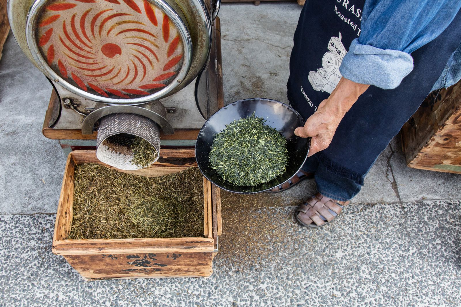 A drum roaster feeding a wooden crate of dark roasted leaf, beside a person in indigo work clothes holding out a black dish of bright green unroasted leaf.