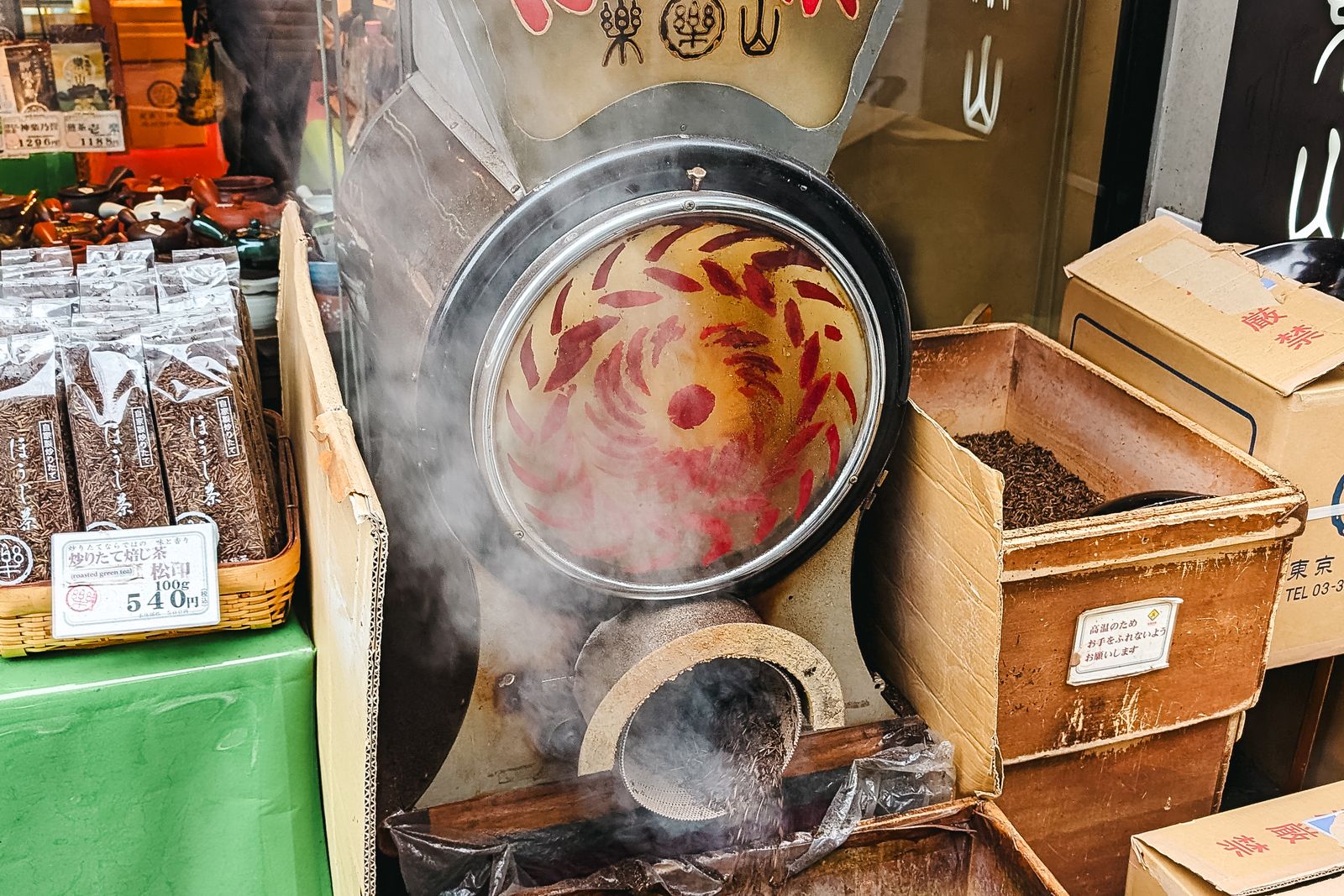 A Japanese tea shopfront with a drum roaster running and steam across the frame, a basket of printed tea packets at left and an open crate of roasted leaf at right.