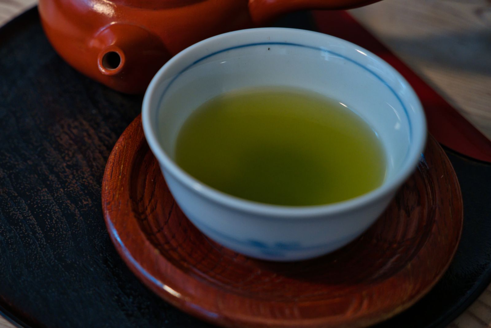 A close overhead view of a white porcelain cup with a blue rim line, holding pale yellow-green brewed tea on a wooden coaster.