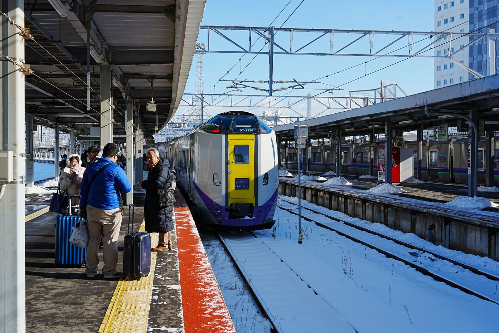 A snow-dusted platform at Hakodate station with an express train arriving and travellers with suitcases at the left