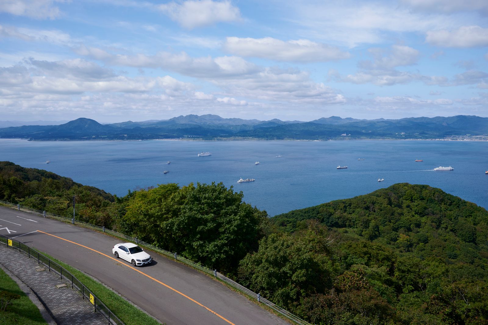 A bay with cargo ships at anchor seen from a road above the coast, blue hills across the water, broken cloud