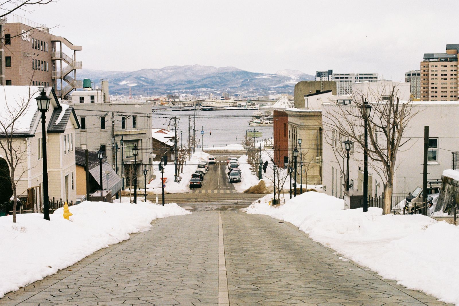 A stone-paved street sloping down between banks of snow to a harbour, with mountains across the water
