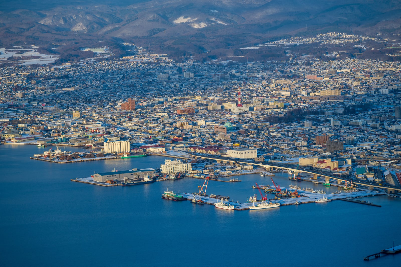 A harbour with moored ships and red cranes seen from above, the city behind and snow on the dark ridge, low sun on the water