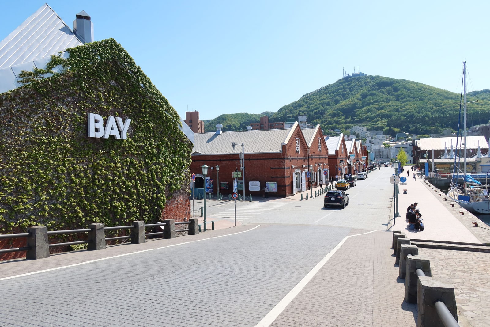 Red-brick warehouses along a paved waterfront road, an ivy-covered brick wall at the left and a green hill behind