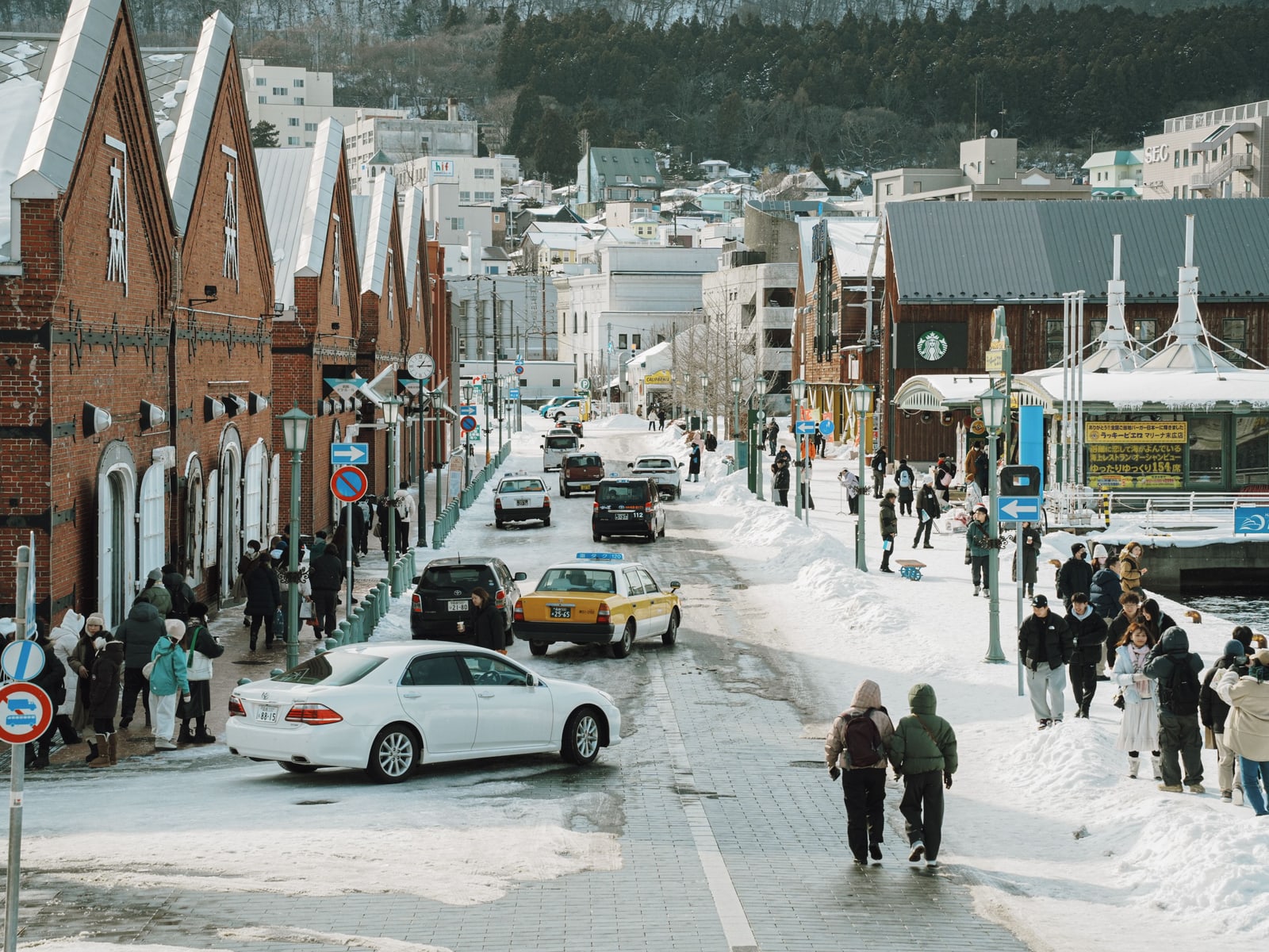 A snowy street beside red-brick gabled warehouses, with taxis, cars and visitors in winter coats
