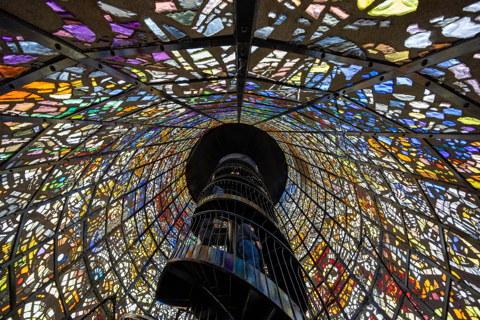 Looking up inside a tower of coloured glass pieces held in a black grid, with a caged spiral staircase and two figures climbing
