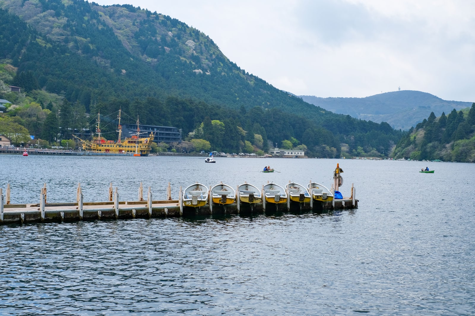 A jetty with six small rowing boats on Lake Ashi, a yellow cruise ship at a pier in the distance, forested hills and an overcast sky