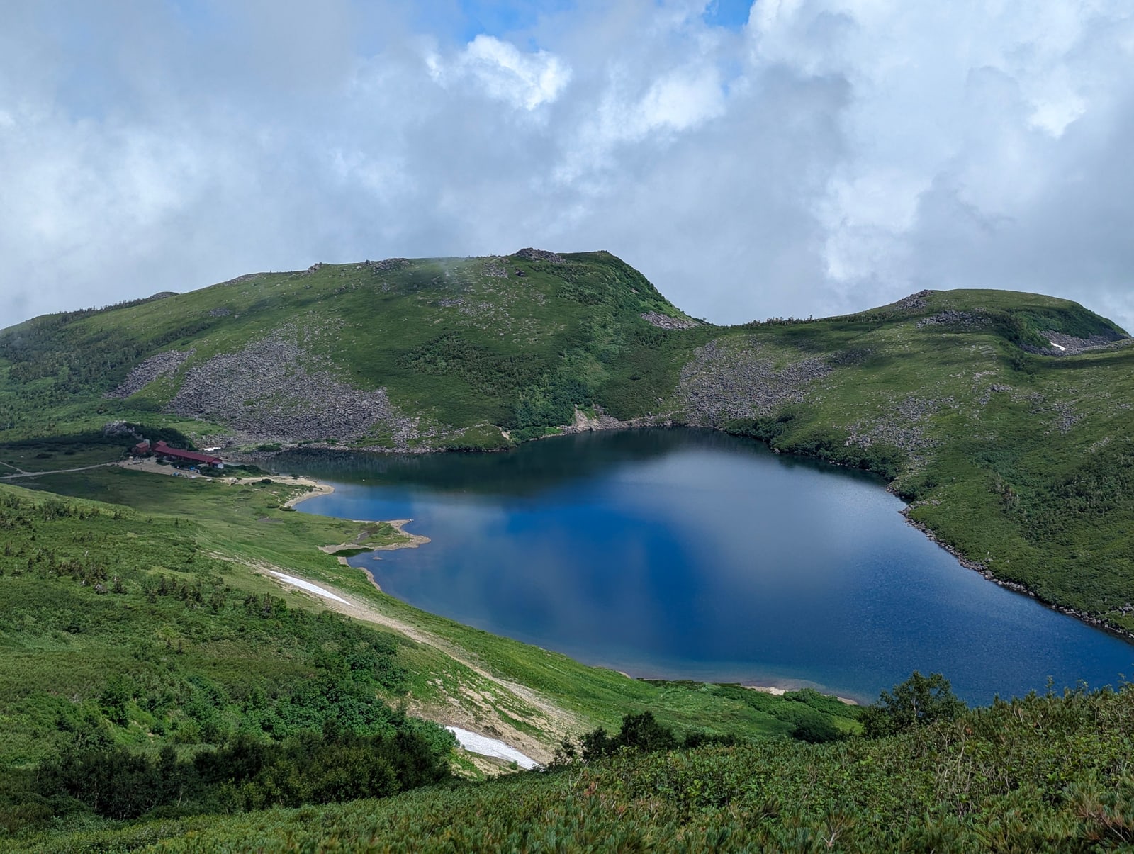 A deep-blue alpine lake in a green basin with a red-roofed mountain hut on the shore, mountains of the Hakuba area in summer