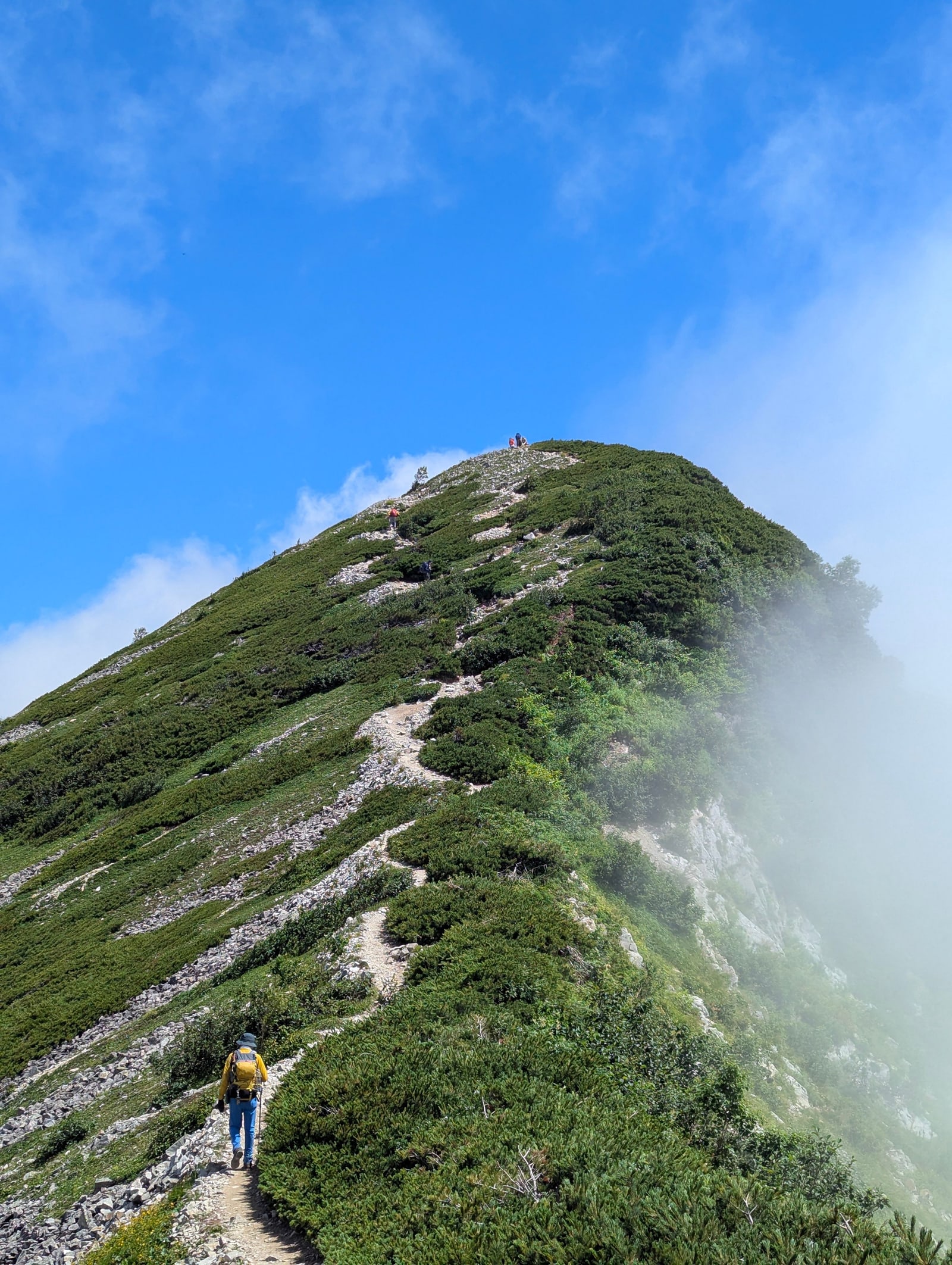 A hiker in a yellow jacket on a stony trail up a green mountain ridge in the Hakuba area, cloud spilling over the side