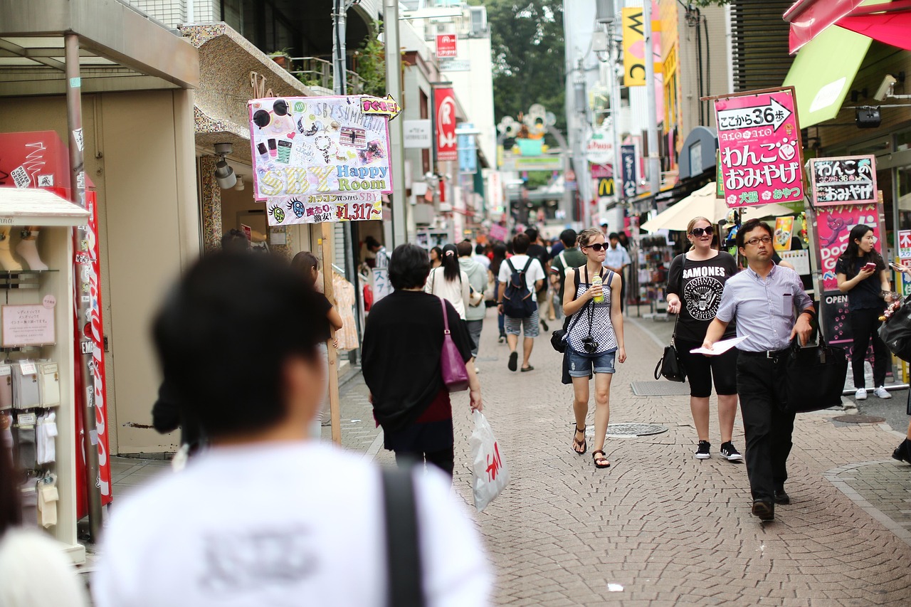A crowded brick-paved shopping street in Harajuku in daylight, with shop signs and sandwich boards on both sides