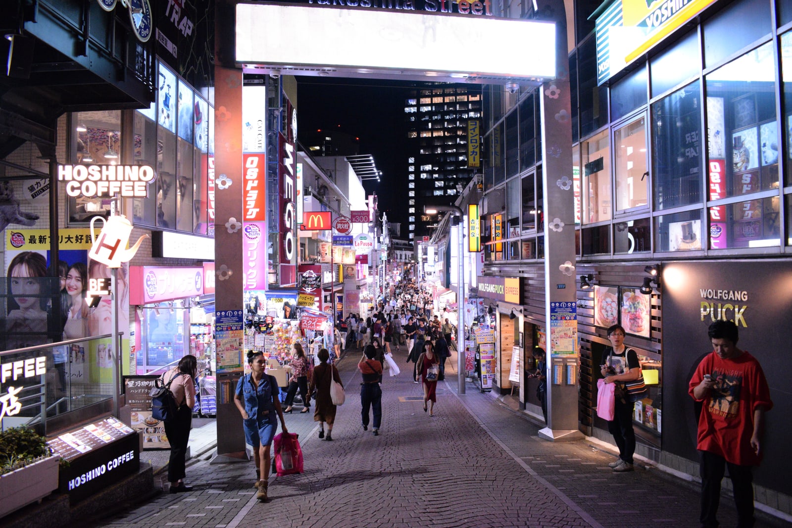 The entrance of Takeshita Street at night under a lit sign, shop signs on both sides and a crowd walking down the cobbled street