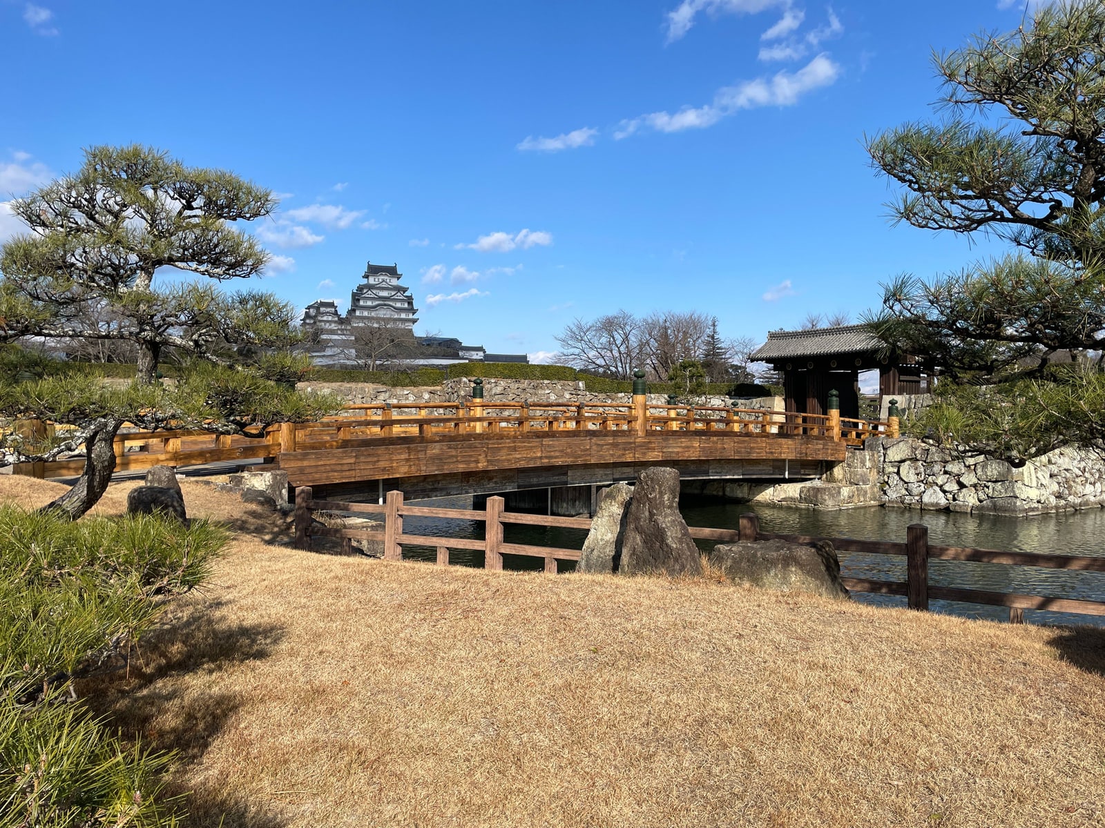 A wooden arched bridge over a pond among pine trees, a small roofed gate to the right and the castle keep small in the distance.