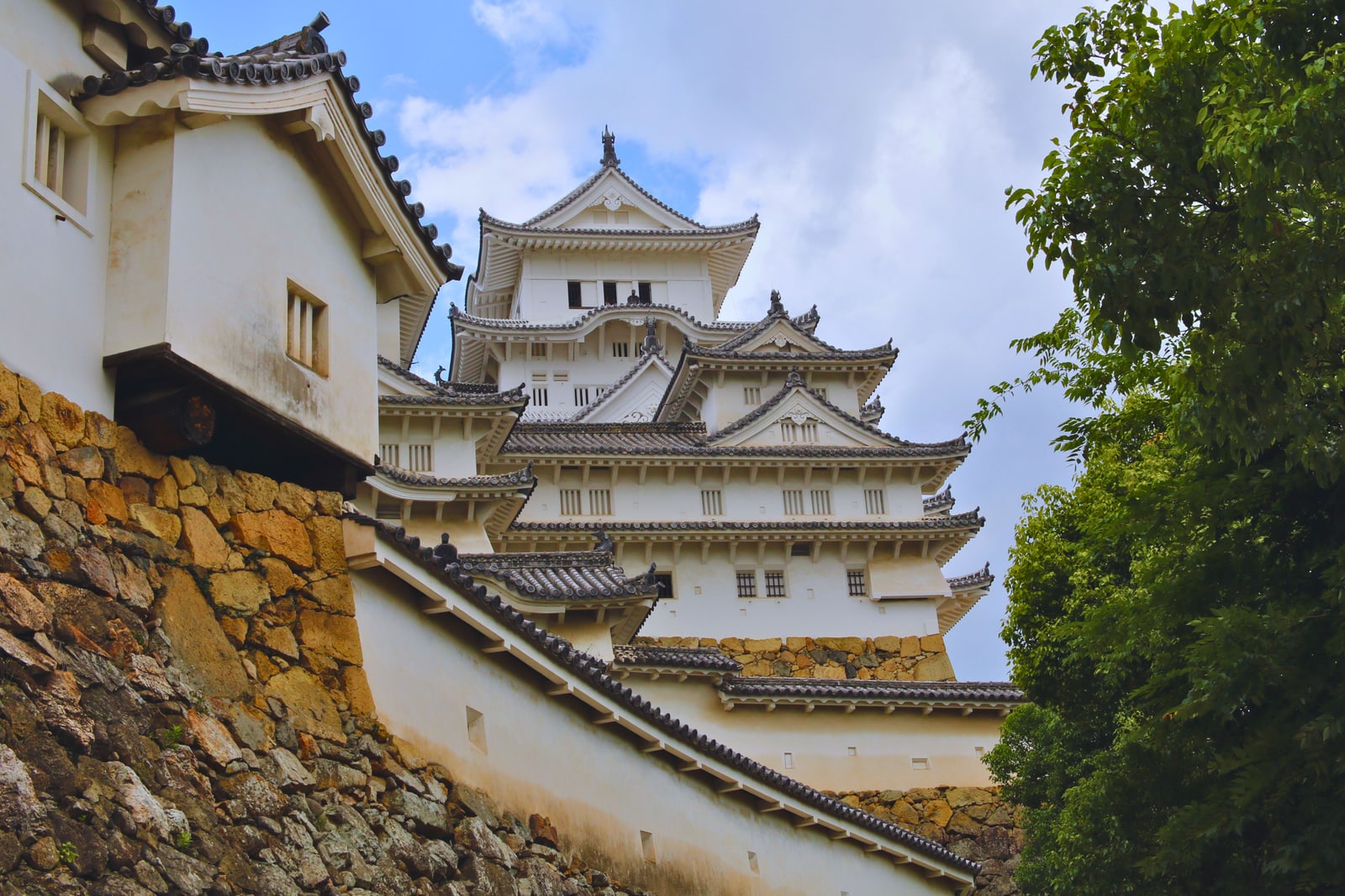 Looking up a sloping stone base and white plastered wall toward the castle keep, a turret close on the left and trees on the right.