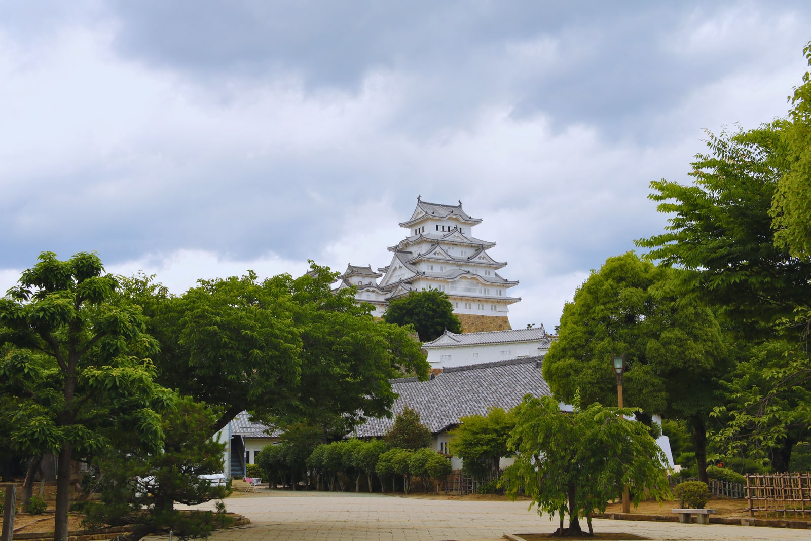 The castle keep rising above green trees and low white walls, a wide paved approach in front under a cloudy sky.
