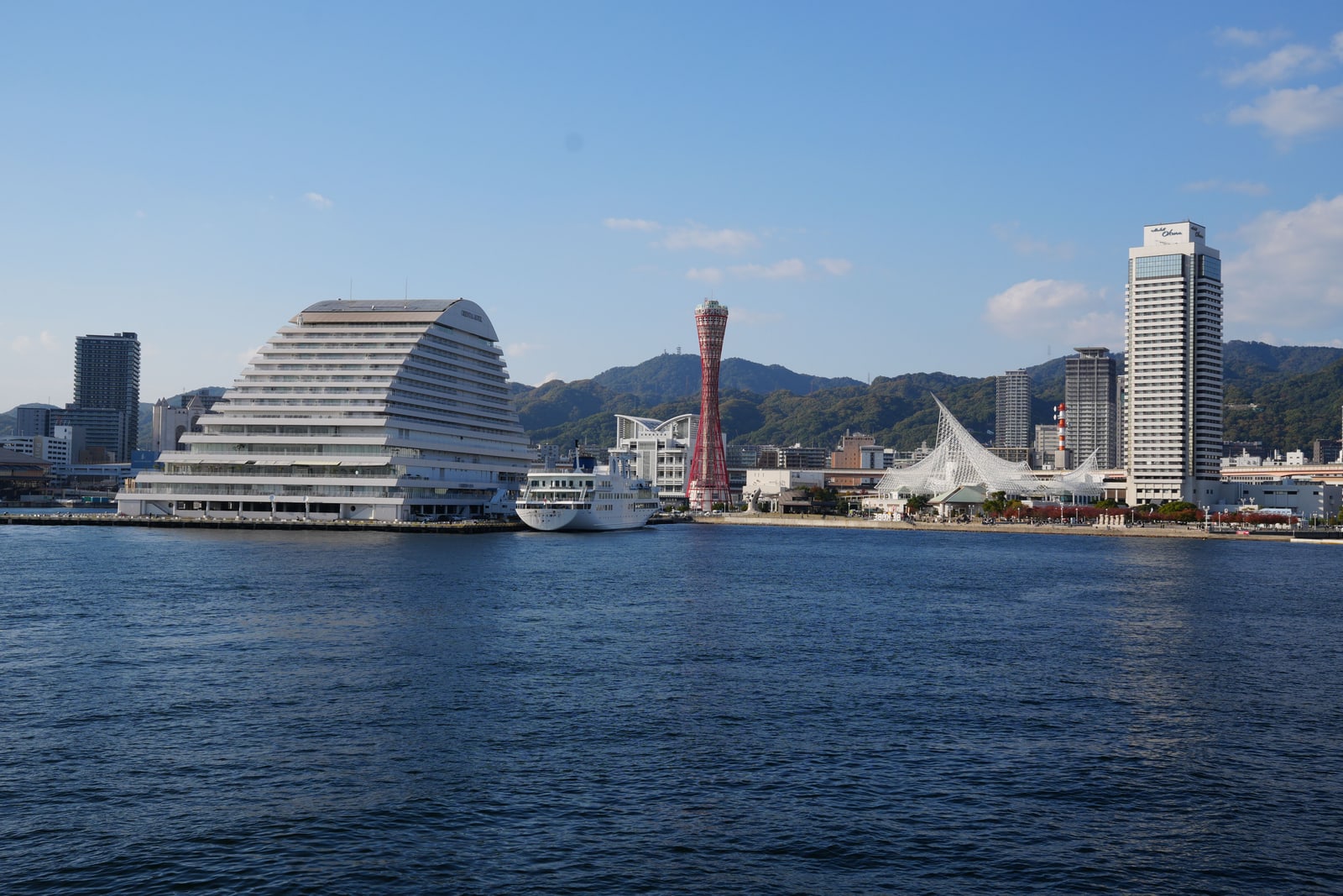 Kobe harbor across blue water, the red lattice Port Tower, a curved white hotel and a tall tower, green hills behind.