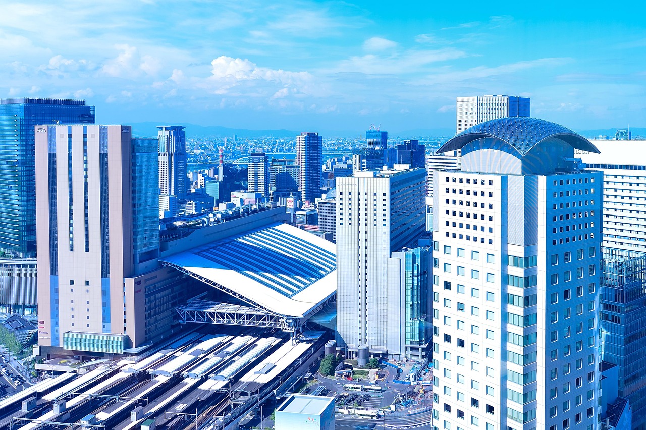 A high view over Osaka Station, its long curved roof over the tracks between office towers under a blue sky.