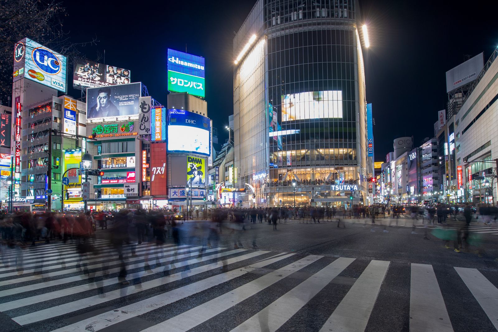 Shibuya Crossing at night in a long exposure, the crowd blurred into streams of motion between the lit signboards — ordinary big-city density, and a preview of what the six avoid-windows do to the whole country's trains, rooms and famous places at once.
