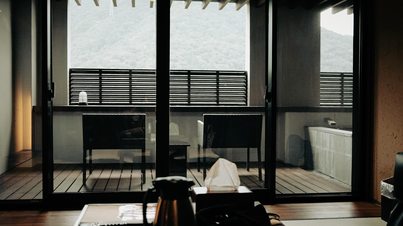 A Hakone inn room looking through glass doors onto a balcony with two chairs and a private outdoor bath, wooded hills beyond.