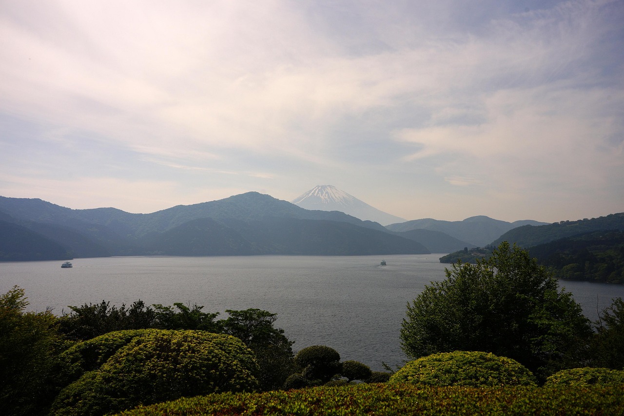 Lake Ashi from a garden above the shore, two sightseeing boats on the water and a snow-capped Mount Fuji beyond the hills.