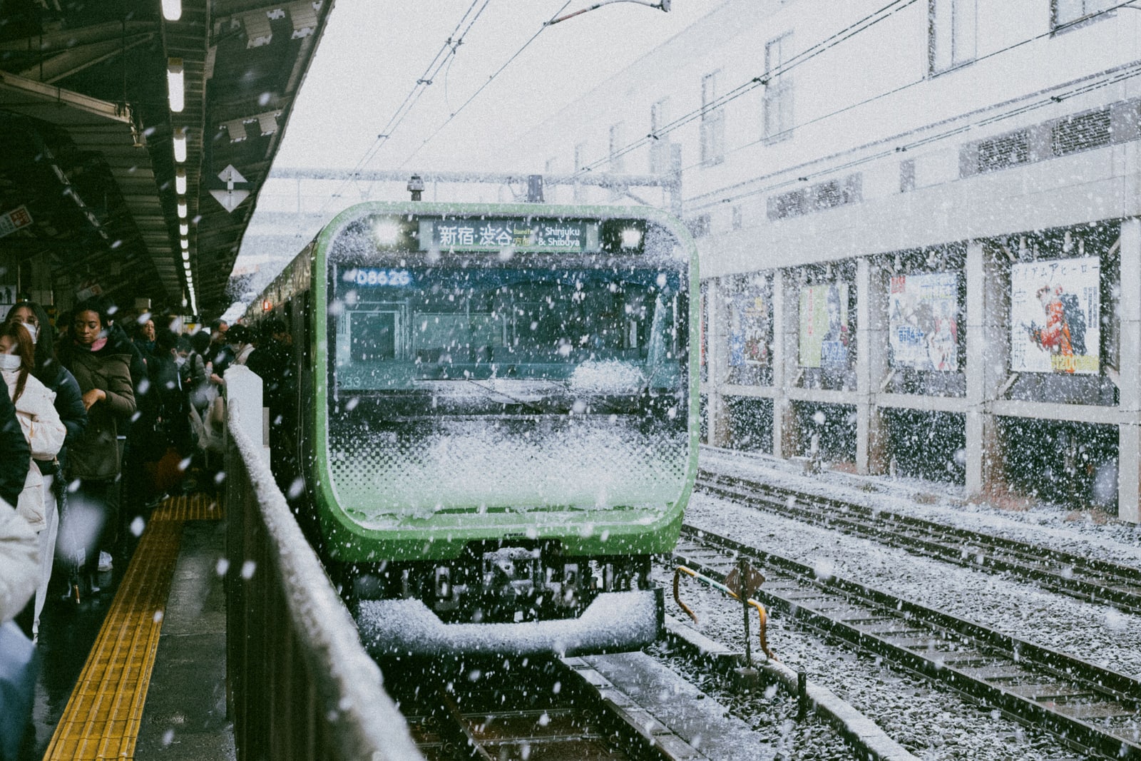 A green-striped commuter train bound for Shinjuku and Shibuya at a crowded platform in falling snow.