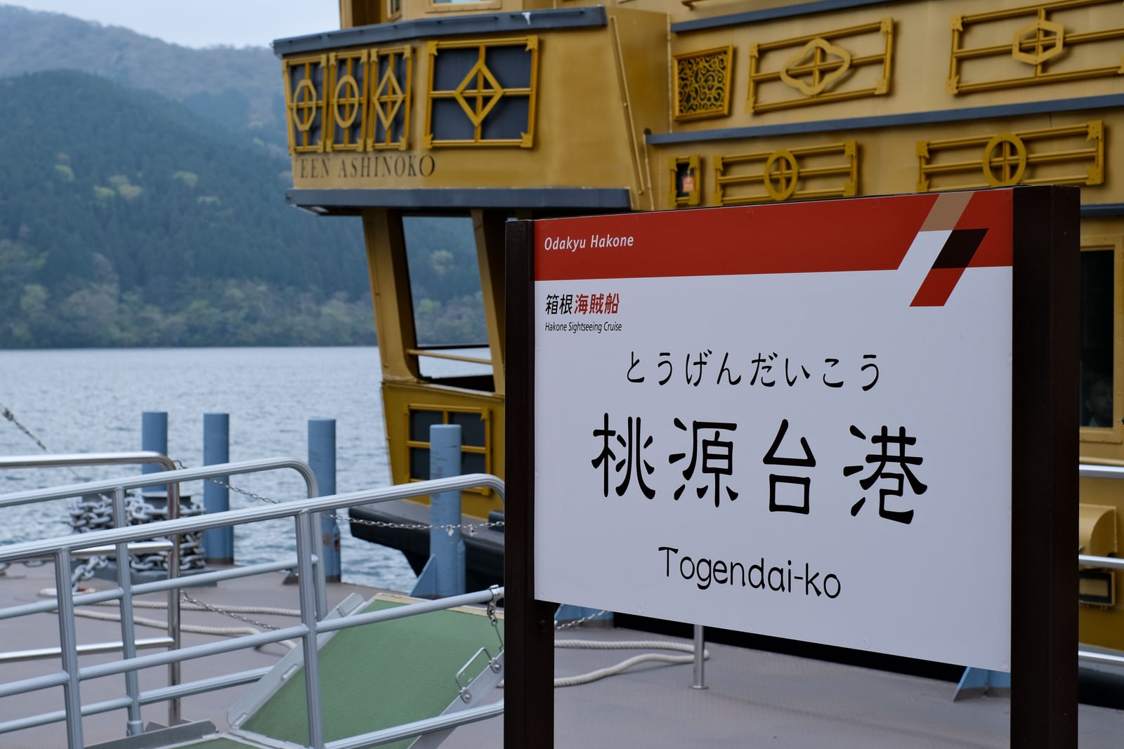Station sign for Togendai-ko port of the Hakone Sightseeing Cruise, a gold-trimmed sightseeing ship moored behind it on Lake Ashi.