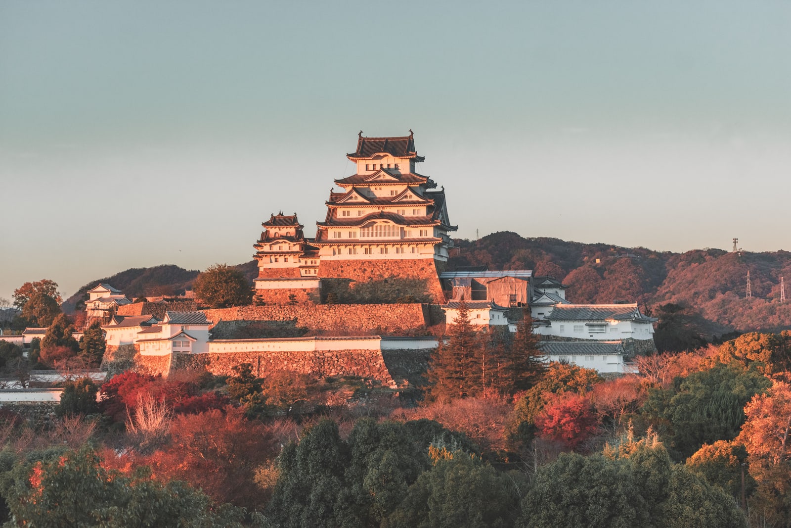 Himeji Castle and its turrets on high stone bases in low orange light, autumn trees below and wooded hills behind