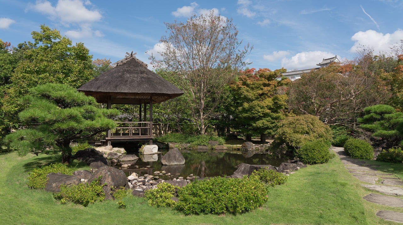 A thatched pavilion beside a small rock-edged pond in a garden lawn, a white tiled-roof building above the trees