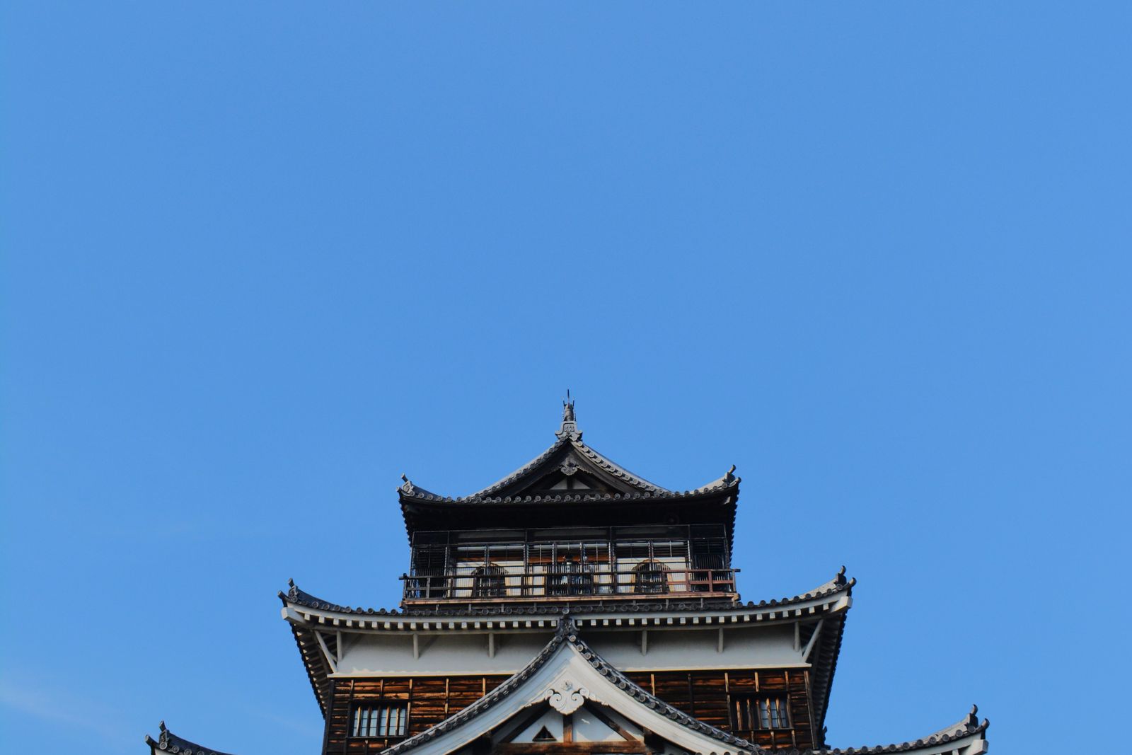 The upper part of Hiroshima Castle's keep — dark plank cladding, white gables, a shachihoko finial — against a clear blue sky.