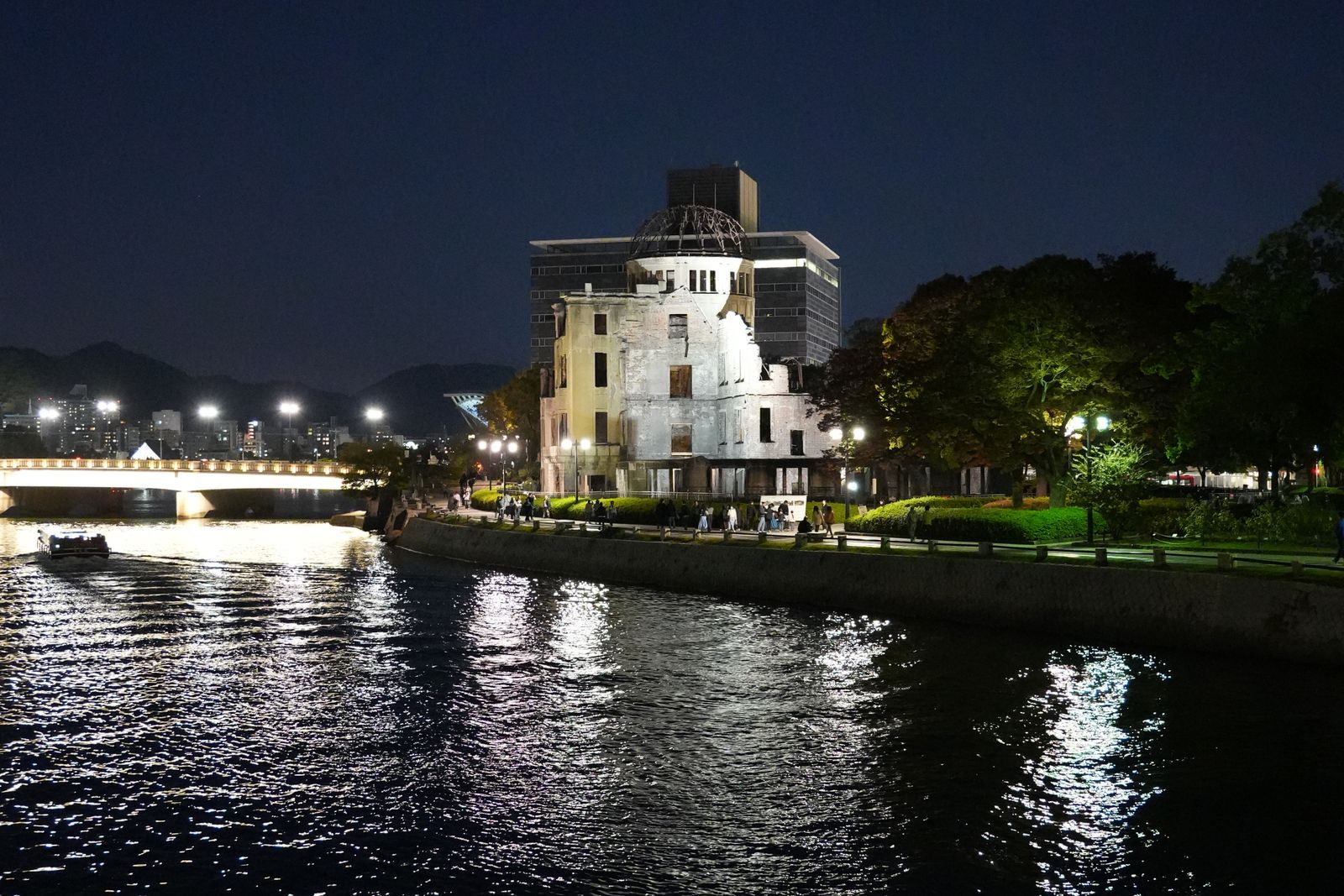 The Atomic Bomb Dome floodlit at night from across the river in Hiroshima, an illuminated bridge at left and promenade lamps along the bank.