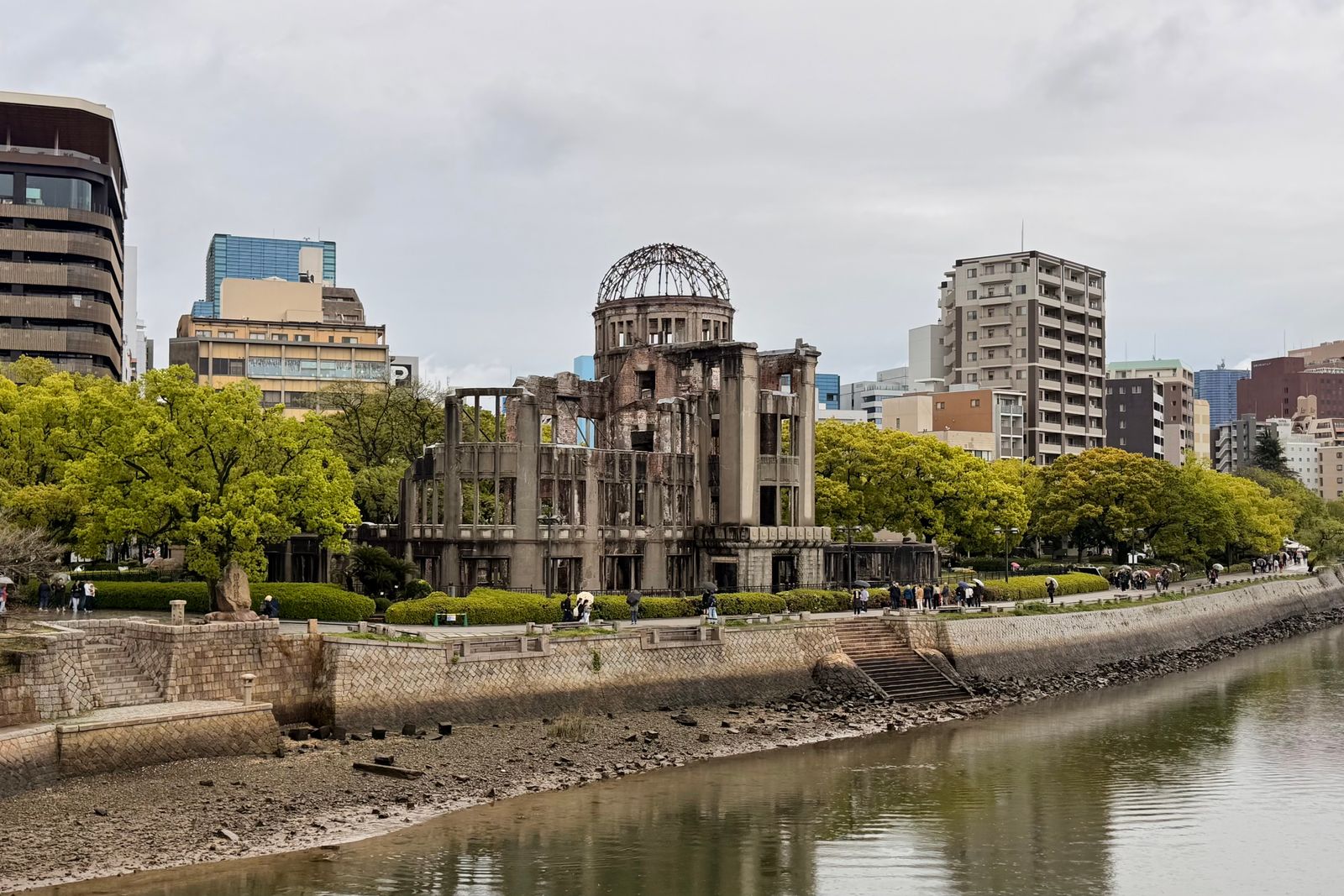 The Atomic Bomb Dome centered across the river in Hiroshima under an overcast sky, fresh spring green on the banks and the riverbed exposed at low tide.
