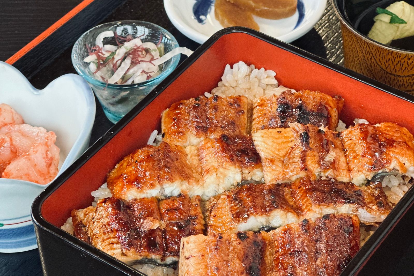 Grilled glazed eel fillets over rice in a red lacquered box with small side dishes on a black tray