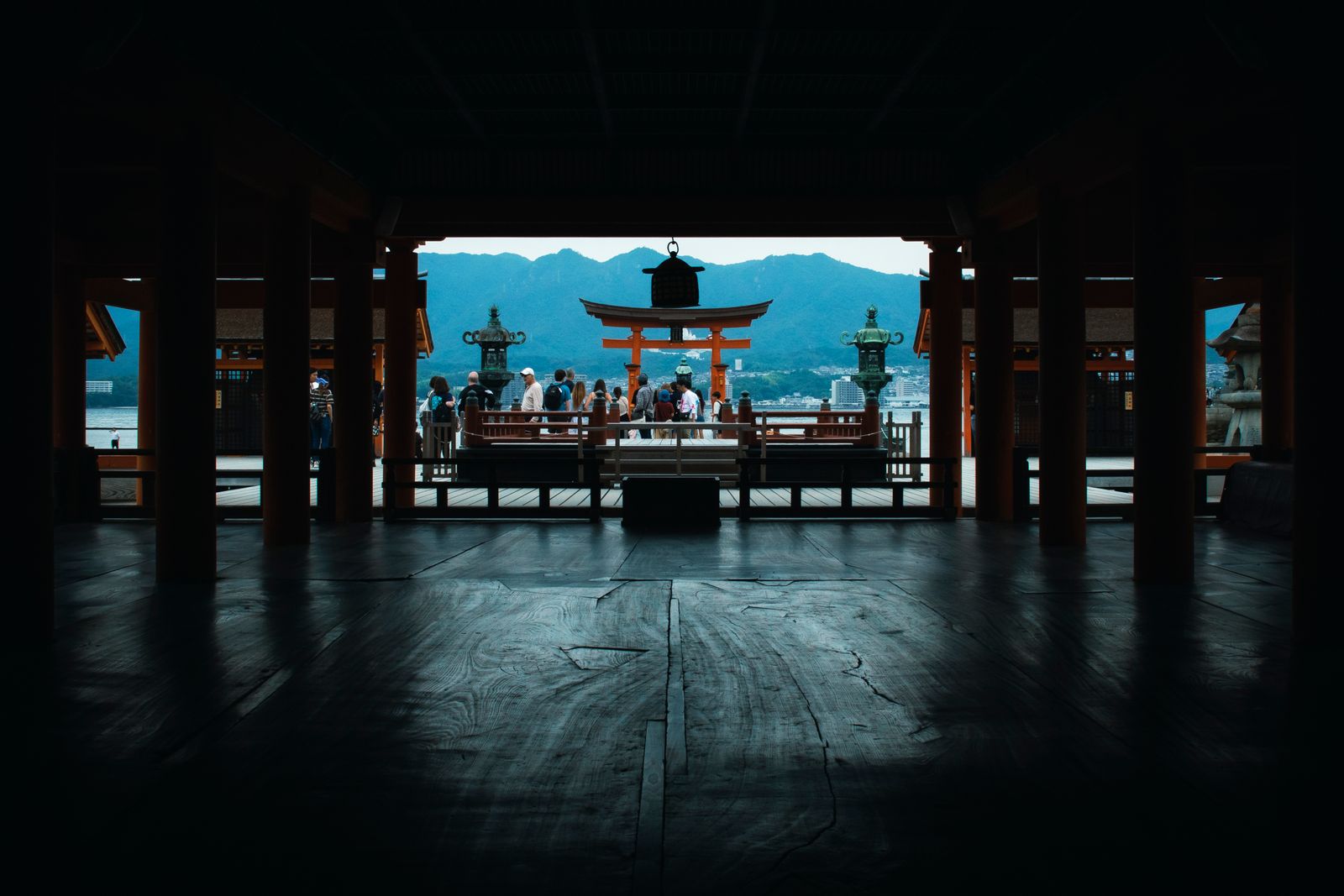 From inside the dark hall of Itsukushima Shrine, looking through vermilion pillars and bronze lanterns across the stage to the great torii and the mountains beyond.