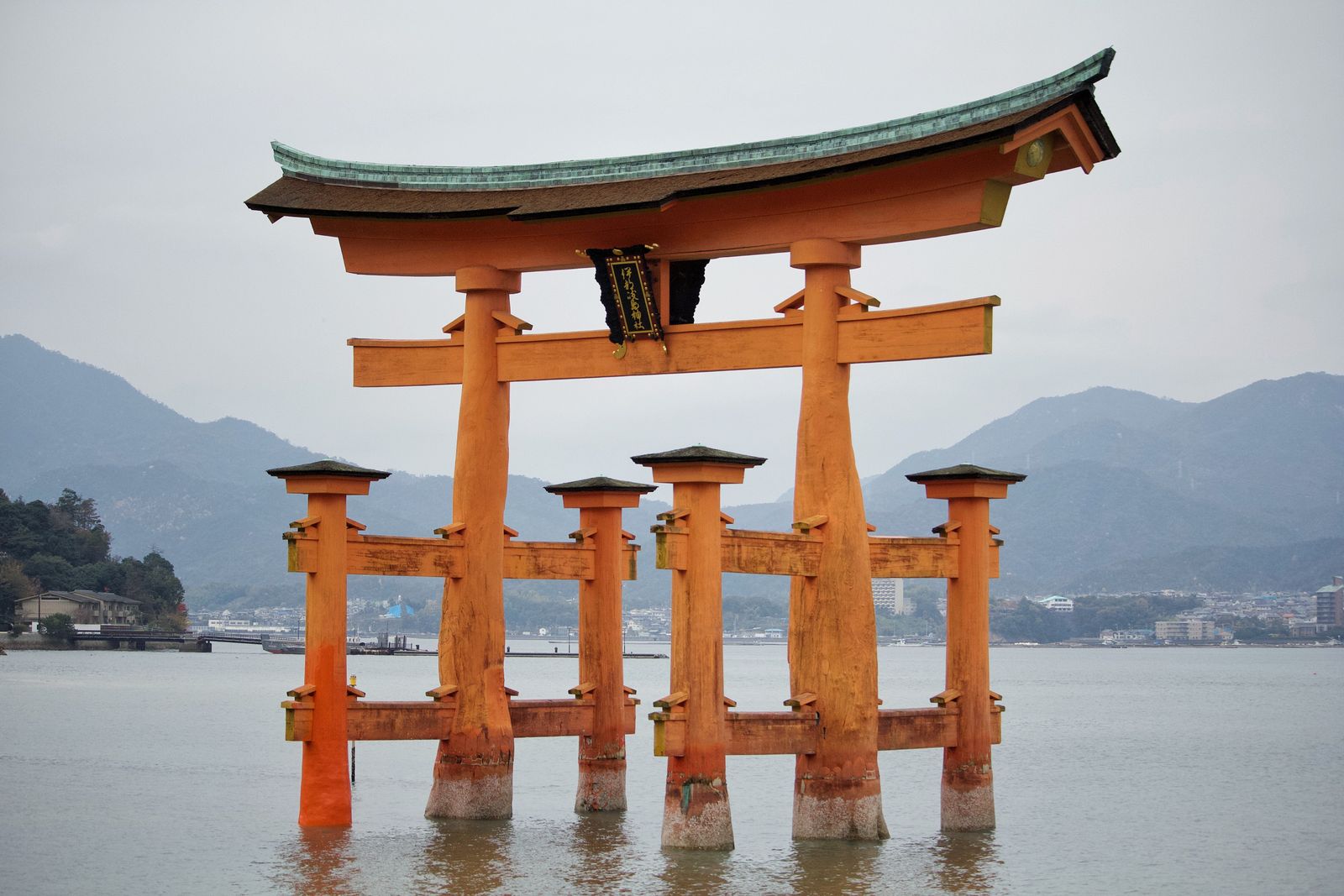 The great torii of Itsukushima Shrine standing in the water, hills and the mainland shore behind under a grey sky.
