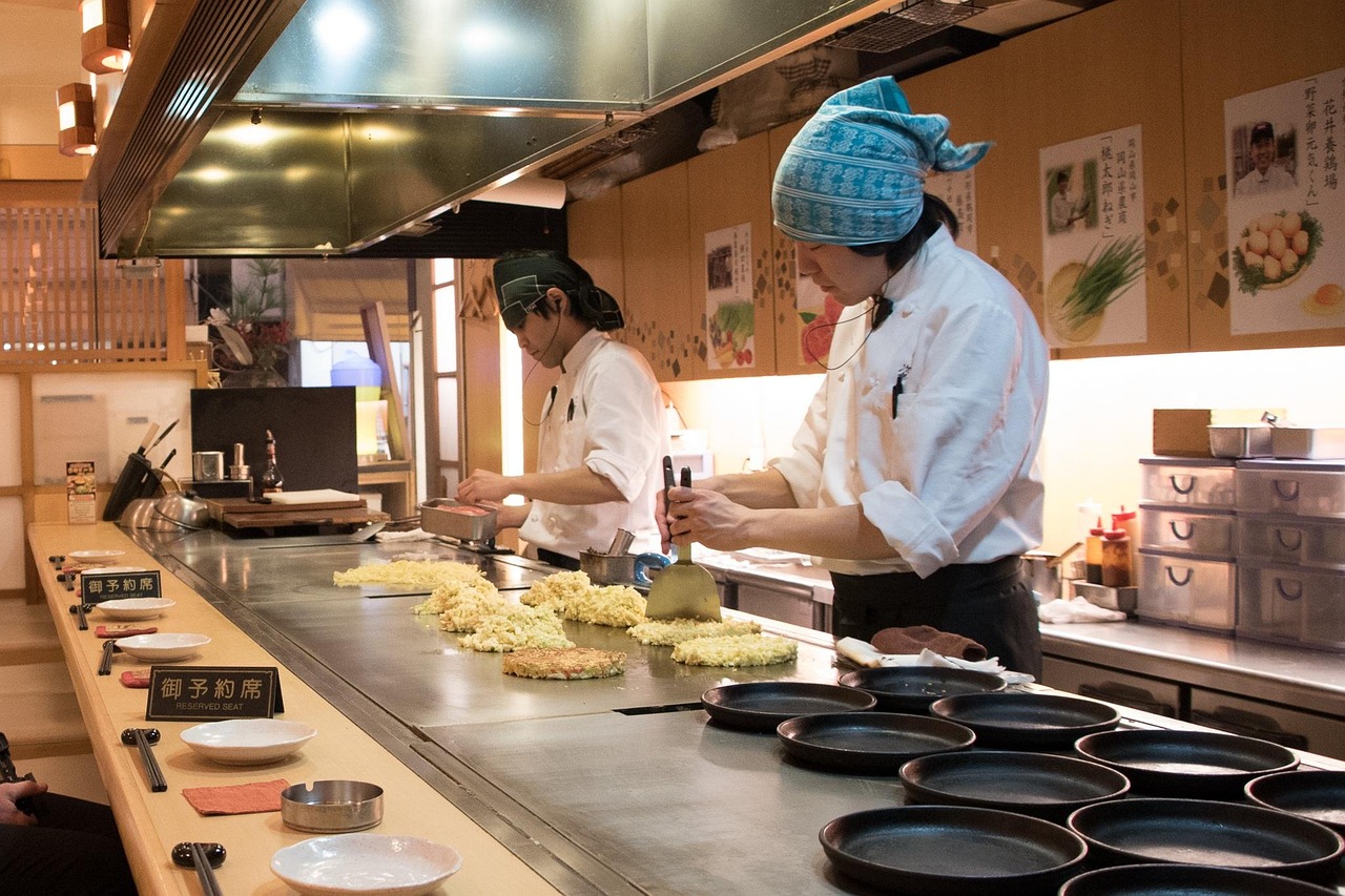 A restaurant counter griddle with two cooks in white: six separate mounds of shredded cabbage laid out on the teppan, and one round at lower left already pressed flat and browning.