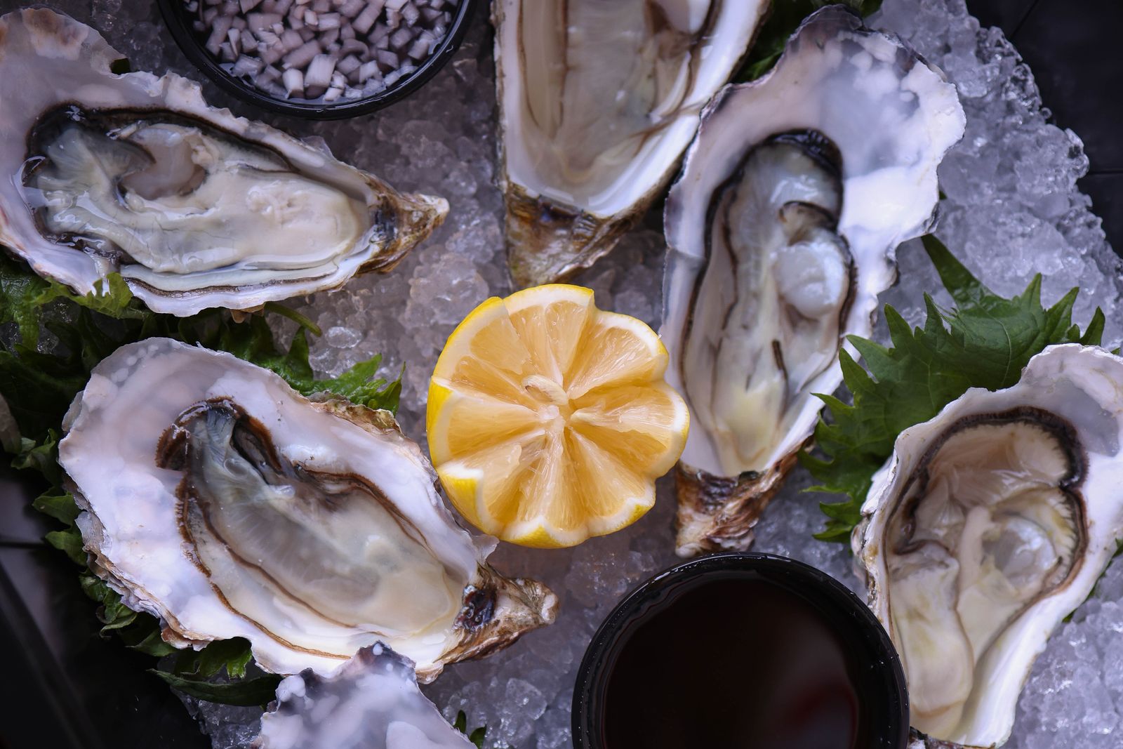Five raw oysters on the half shell on crushed ice, a lemon cut into a flower at the center, shiso leaves under the shells, a small bowl of dark dipping sauce and a bowl of chopped shallot.