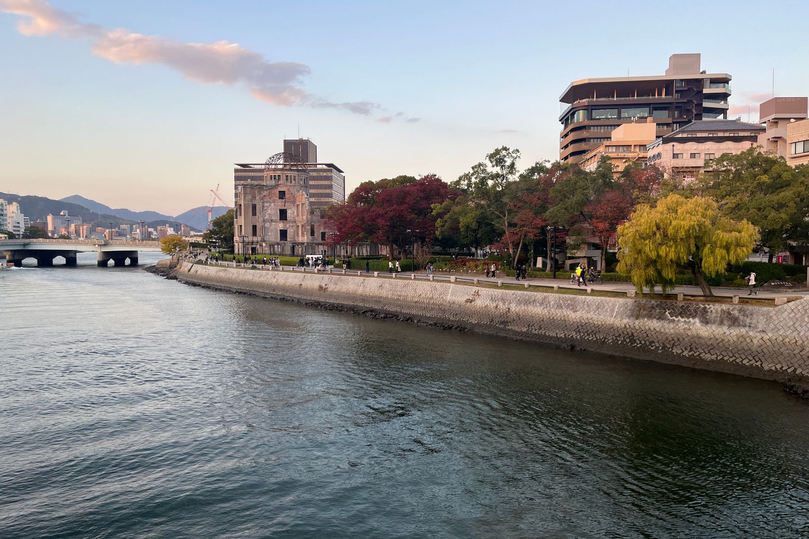 The Atomic Bomb Dome across the river in Hiroshima, autumn-red maples and a willow on the near bank, a bridge at left, hills behind in soft light.