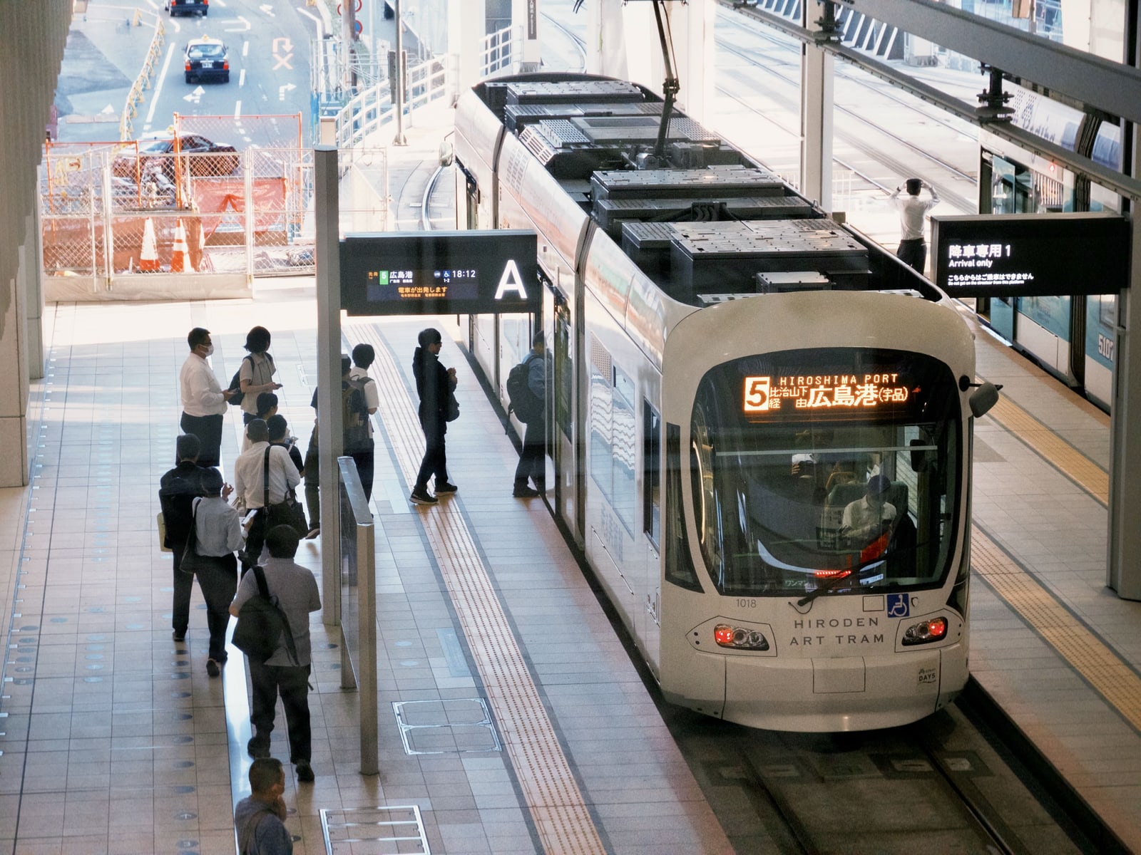 A white Hiroden tram with a route 5 destination board for Hiroshima Port (Ujina) at an elevated platform, passengers waiting at left