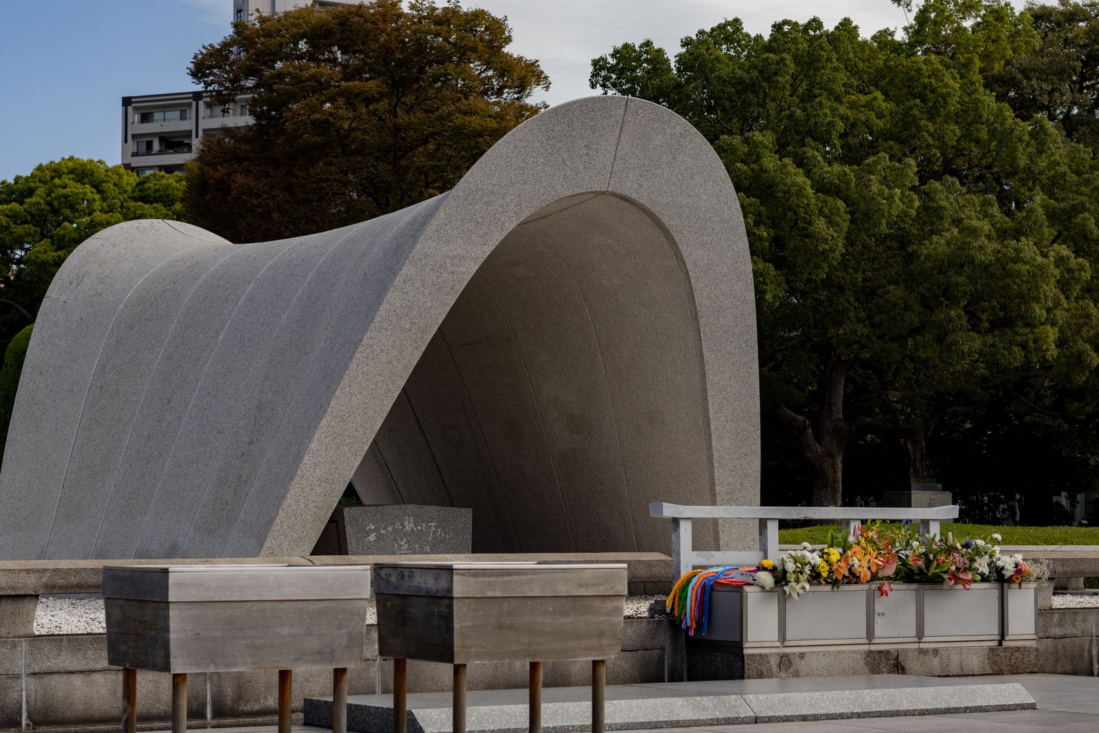 The saddle-shaped granite Cenotaph in Peace Memorial Park seen from the side, wooden offering boxes and a planter of cut flowers in front, trees behind