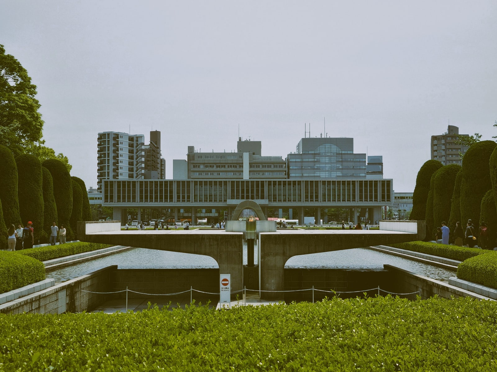 A long raised museum building seen across a pond in Peace Memorial Park, a small flame on a concrete stand in front and clipped hedges at both sides under an overcast sky