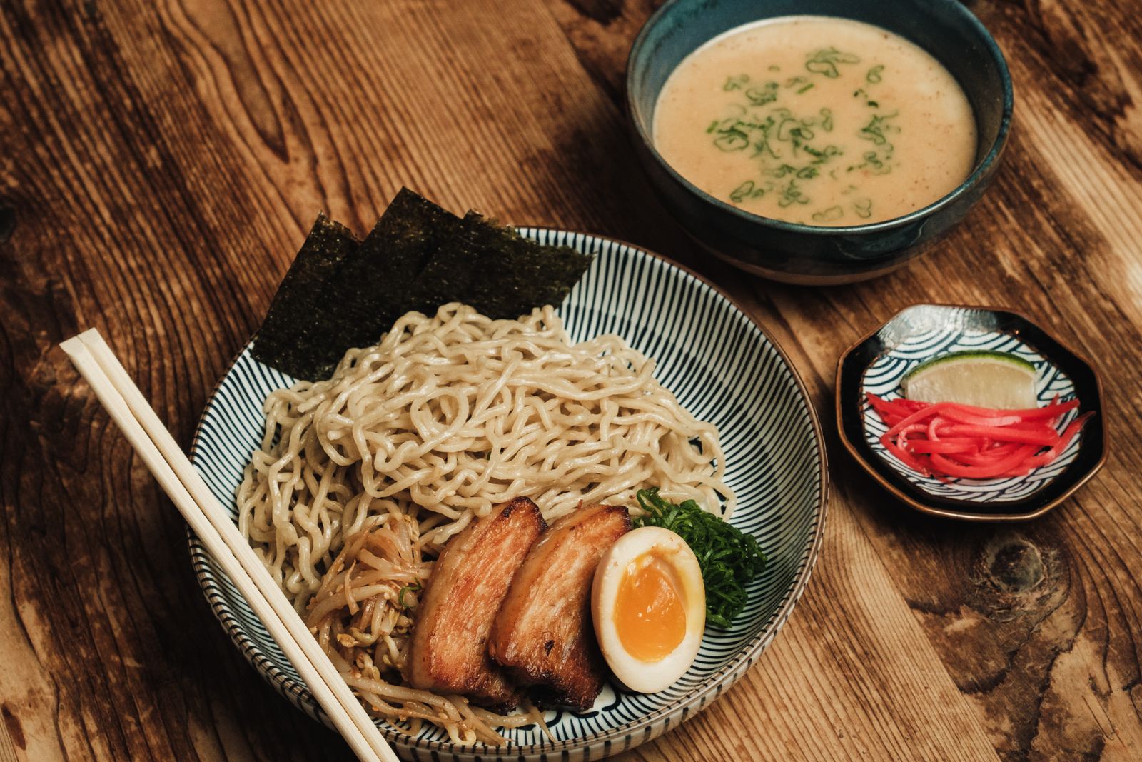 A bowl of tsukemen noodles topped with grilled pork, a soft-boiled egg, nori and bean sprouts, chopsticks resting on the rim, with a bowl of dipping broth beside it.