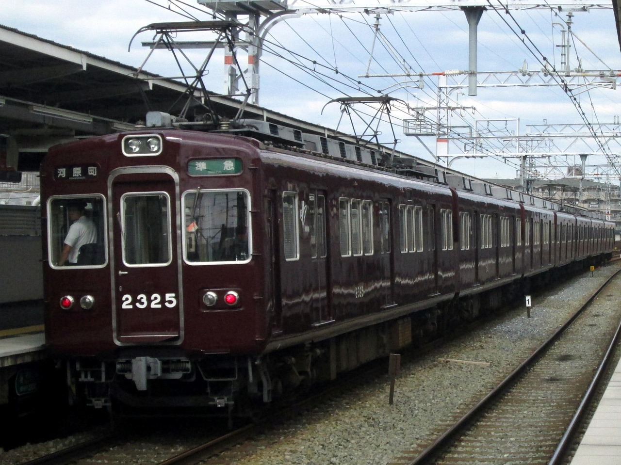A maroon Hankyu train, car 2325, standing at an open-air platform under overhead wires, the driver visible in the cab.