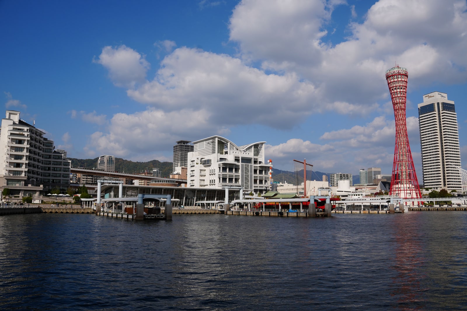 Kobe harbor across open water, the red lattice Port Tower beside a tall pale hotel, blue sky with cloud.