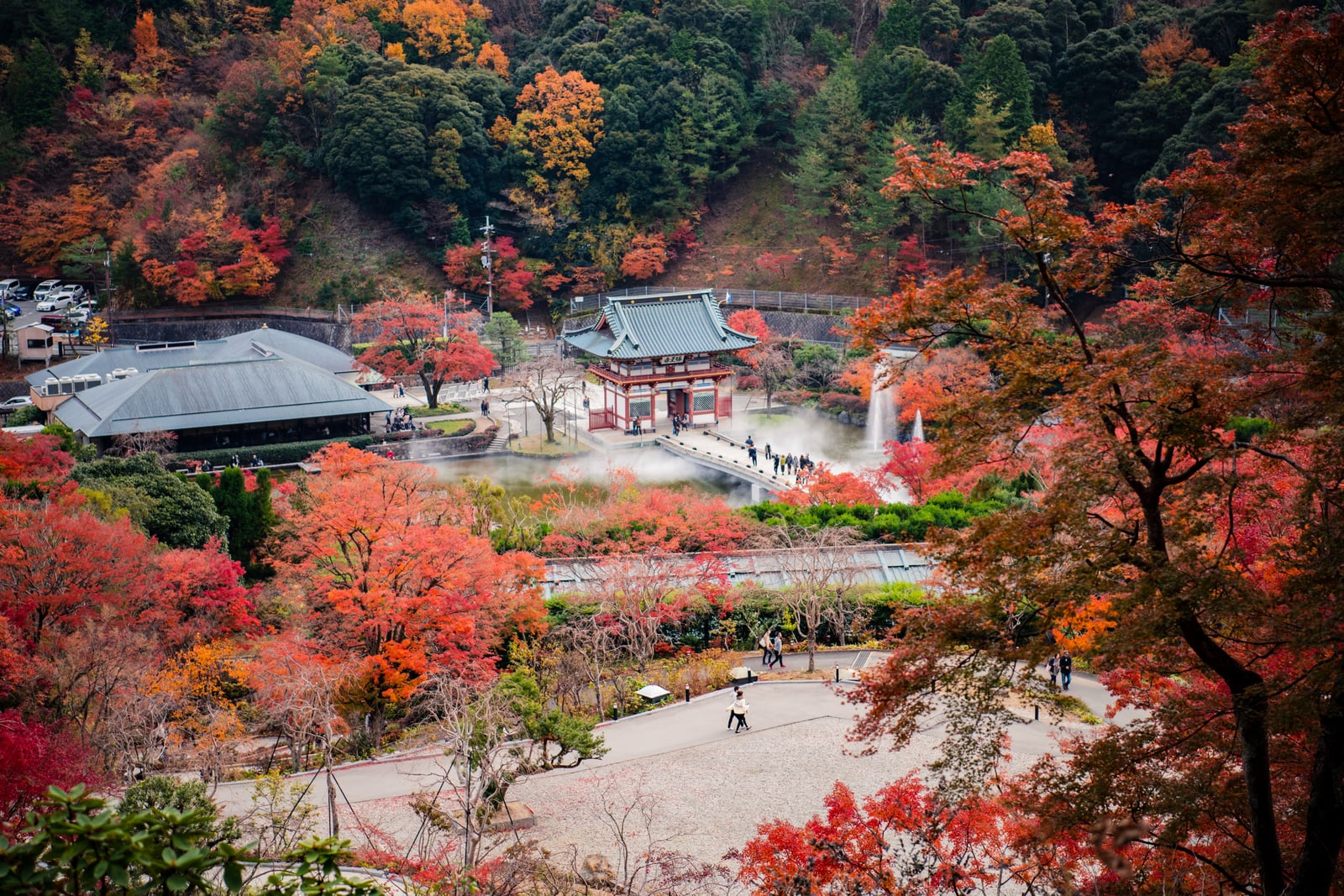 A wooded valley in full autumn red and orange, a small red-pillared hall at the foot of the slope and walkers on a paved path.