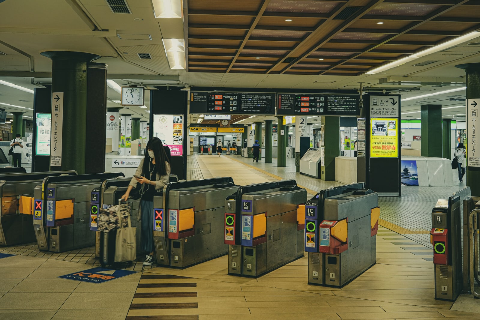 A row of automatic ticket gates in a station concourse, one traveler with a suitcase at a gate, departure boards overhead.