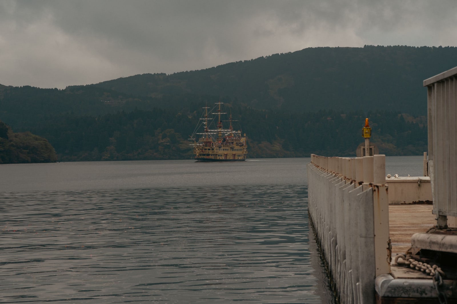 A sightseeing ship on gray water under low cloud, forested hills behind and a wooden pier in the foreground.